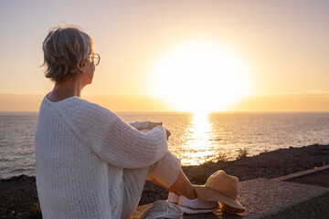 Back view of relaxed and romantic senior woman or pensioner sitting admiring the sea at sunset...