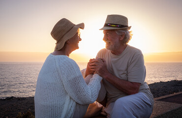 Happy and romantic caucasian couple hands in the hands sitting outdoors at sea at sunset light looking in the eyes - old seniors or pensioners enjoying vacations together