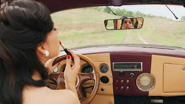 Young attractive retro woman looking in rear view mirror painting her red lips doing applying make up. Pin up hairstyle long hair. Beautiful pin up girl, smiling face Distracted driver. Sexy lady back