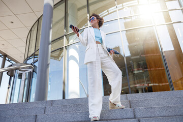 Attractive young businesswoman in white suit holding phone standing near business centre