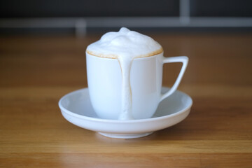 white cup of cappuccino with flowing foam on a wooden table against a black tile wall