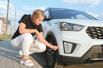 Deurstickers Woman squats down in front of her car and looks at a scratch on the bumper. An attractive woman with blond hair inspects a damaged car. © Galina