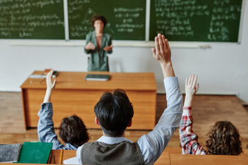 Rear view of several youthful students keeping their hands raised while sitting by desks in front of teacher explaining ne wsubject at lesson