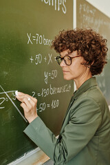 Young confident teacher in eyeglasses and smart casualwear drawing angle on blackboard with chalk...