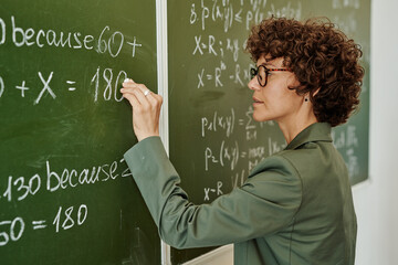 Side view of young teacher with short dark curly hair writing down equation on blackboard while...