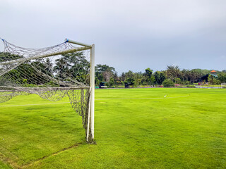 View through the net of football gates to a large football field with a green lawn, a gate opposite and trees. Sports, team sports
