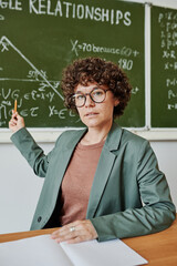 Confident teacher of geometry pointing at drawing of triangle on blackboard while explaining new...