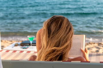 Young blonde woman reading a book and relaxing on a daybed iat the beach