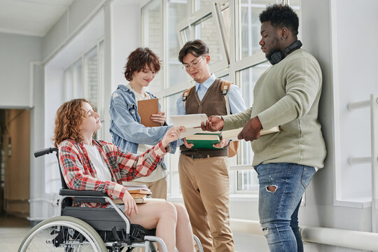 Cute teenage girl with disability taking copybook of African American guy standing in front of her while their classmates having discussion