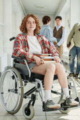 Fototapeta na wymiar Cute teenage female student in wheelchair sitting in front of camera in college corridor against group of her classmates talking at break