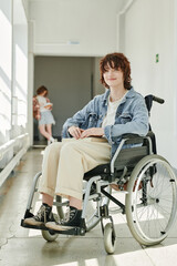Fototapeta na wymiar Happy teenage student with physical disability sitting in wheelchair in college corridor and looking at camera against her classmate
