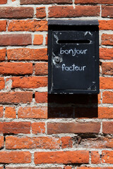 Älterer schwarzer Briefkasten an einer roten Ziegelsteinmauer, auf dem bonjour facteur geschrieben steht, vertikal