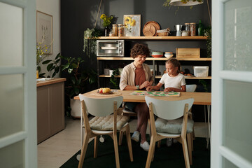 Young woman and her youthful daughter playing board game togather while sitting by wooden kitchen table at leisure