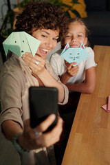 Young cheerful woman and her cute youthful daughter with origami toys looking at smartphone camera while making selfie by table - obrazy, fototapety, plakaty