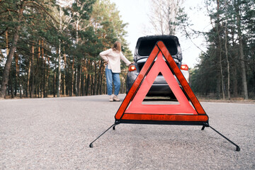 Red emergency triangle with blurred car and woman calling car mechanic in the background. Broken car on the road