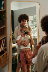 Reflection in mirror of young woman embracing her cute youthful daughter with photocamera taking shot with mother