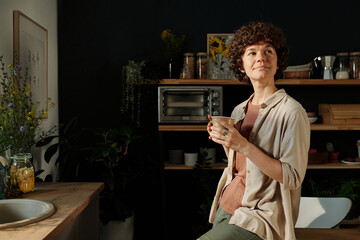 Young mindful brunette woman in casualwear holding cup of tea or coffee while sitting on kitchen...