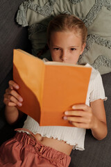 Cute youthful schoolgirl in casualwear keeping head on pillow while relaxing on couch and reading interesting book in orange cover