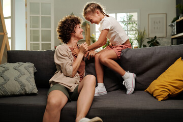 Cute cheerful girl and her happy mother laughing at something funny while relaxing on couch and...