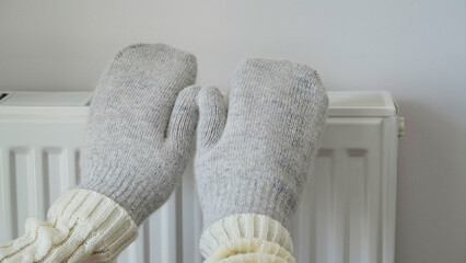 Close-up of woman in white sweater and gray mittens warms her hands over a radiator against a white wall. Macro photo of female hands, copy space. Gas saving concept in winter. Gas crisis in Europe.