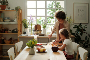 Young woman making sandwiches for youthful daughter and herself for breakfast while standing by kitchen table next to girl eating - obrazy, fototapety, plakaty