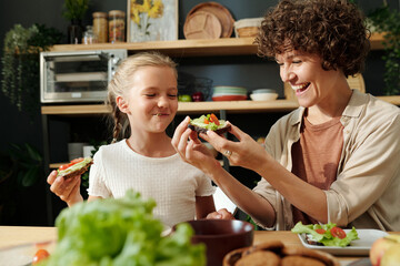 Young laughing brunette woman looking at vegetable sandwich in her hands while sitting by served table next to her youthful daughter