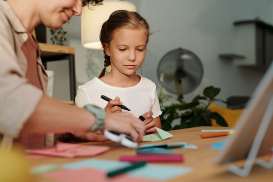 Cute girl looking at her mother folding paper while creating origami at leisure after watching online course or masterclass - Powered by Adobe