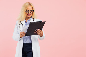Mature woman doctor in glasses with stethoscope holding folder on the pink background with copy space