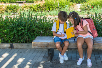 Elementary age children doing school exercises sitting on a wooden bench in the schoolyard. Girl and boy with backpack and school supplies. Education. Back to school.Lifestyle.Children with backpacks