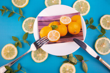 Fresh yellow lemon slices and green mint leaves on blue background around of plate with printed photo, fork, knife. Flat lay, top view. Citrus fruits concept