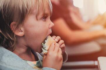 little candid kid boy five years old eats burger or sandwich food sitting in airplane seat on flight traveling from airport. children take a bite. child in air plane eating lunch or dinner meal. flare