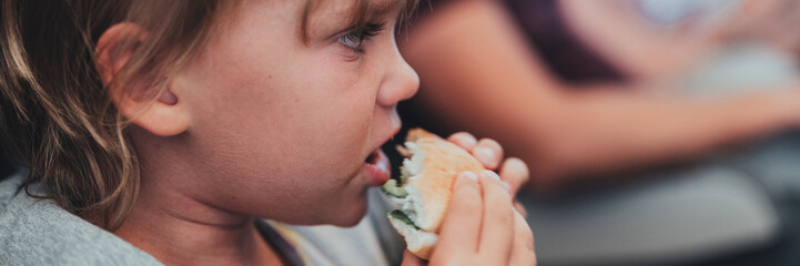 little candid kid boy five year old eats burger or sandwich food sitting in airplane seat on flight traveling from airport. children take a bite. child in air plane eating lunch or dinner meal. banner