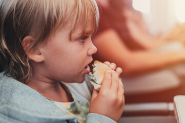 little candid kid boy five years old eats burger or sandwich food sitting in airplane seat on flight traveling from airport. children take a bite. child in air plane eating lunch or dinner meal. flare