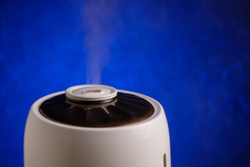 Modern humidifier generates vapor on blue background. Steam from working air purifier.