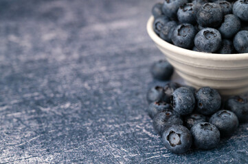 Close-up of a blueberries in a white bowl surrounded by blueberry berries on a dark blue metallic background.