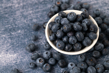 Close-up of a blueberries in a white bowl surrounded by blueberry berries on a dark blue metallic background.