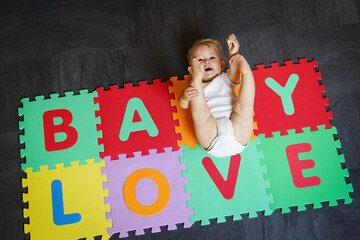 bird view of colorful kids puzzle mat playground in nursery with letters like Baby Love written on them lying on the floor while one year old blond baby in white body is playing
