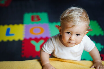 bird view of colorful kids puzzle mat playground in nursery with letters like Baby Love written on them lying on the floor while one year old blond baby in white body is playing
