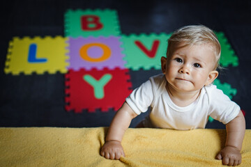 bird view of colorful kids puzzle mat playground in nursery with letters like Baby Love written on them lying on the floor while one year old blond baby in white body is playing
