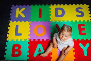 bird view of colorful kids puzzle mat playground in nursery or at home with the letters KIDS LOVE BABY on it lying on the floor while one year old blond baby is sitting there in a grumpy sad mood