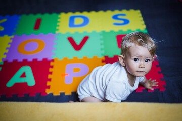 Fototapeta na wymiar bird view of colorful kids puzzle mat playground in nursery with letters like Baby Love written on them lying on the floor while one year old blond baby in white body is playing 