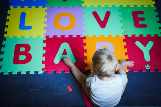bird view of colorful kids puzzle mat playground in nursery with letters like Baby Love written on them lying on the floor while one year old blond baby in white body is playing

