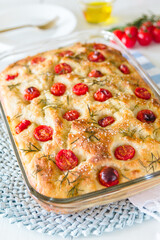 Tomato and rosemary focaccia in a glass baking tray and on a blue table set and white wooden surface, tomatoes and olive oil bottle with white plate and fork