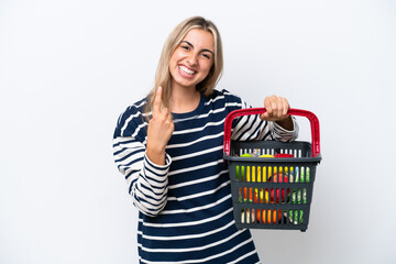 Young caucasian woman holding a shopping basket full of food isolated on white background doing coming gesture