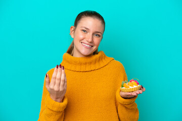 Young caucasian woman holding a tartlet isolated on blue background inviting to come with hand. Happy that you came