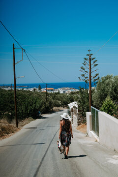 The young woman walks the quiet streets of the picturesque city of Hersonissos in Greece. She wears a white hat and a colorful dress. In the background you can see on the horizon, the turquoise water.