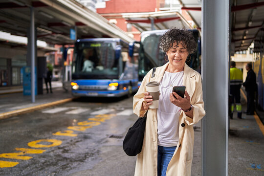 A Mature Woman In A Bus Terminal Using Her Phone And Holding Cup Of Coffee