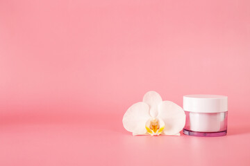 Glass cosmetic jar with cream with orchid flower isolated on pink background.