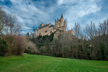 Fototapeta na wymiar Views of the Alcazar of Segovia with the deciduous trees at the base of the cliff and a field of green grass
