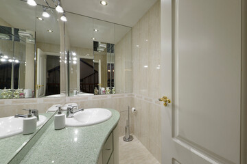 Bathroom with white porcelain sink on light green stone countertop, large frameless wall mirror, wooden cabinet with drawers and white lacquered door